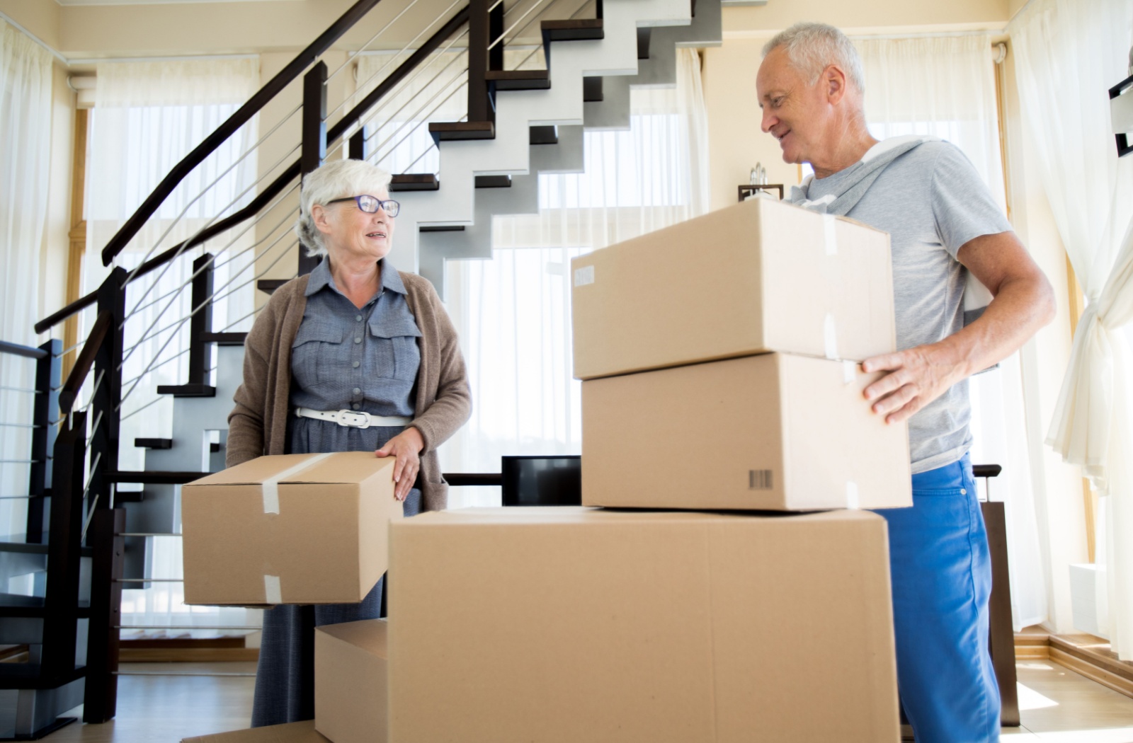 An older adult woman and an older adult man smiling while carrying cardboard boxes.