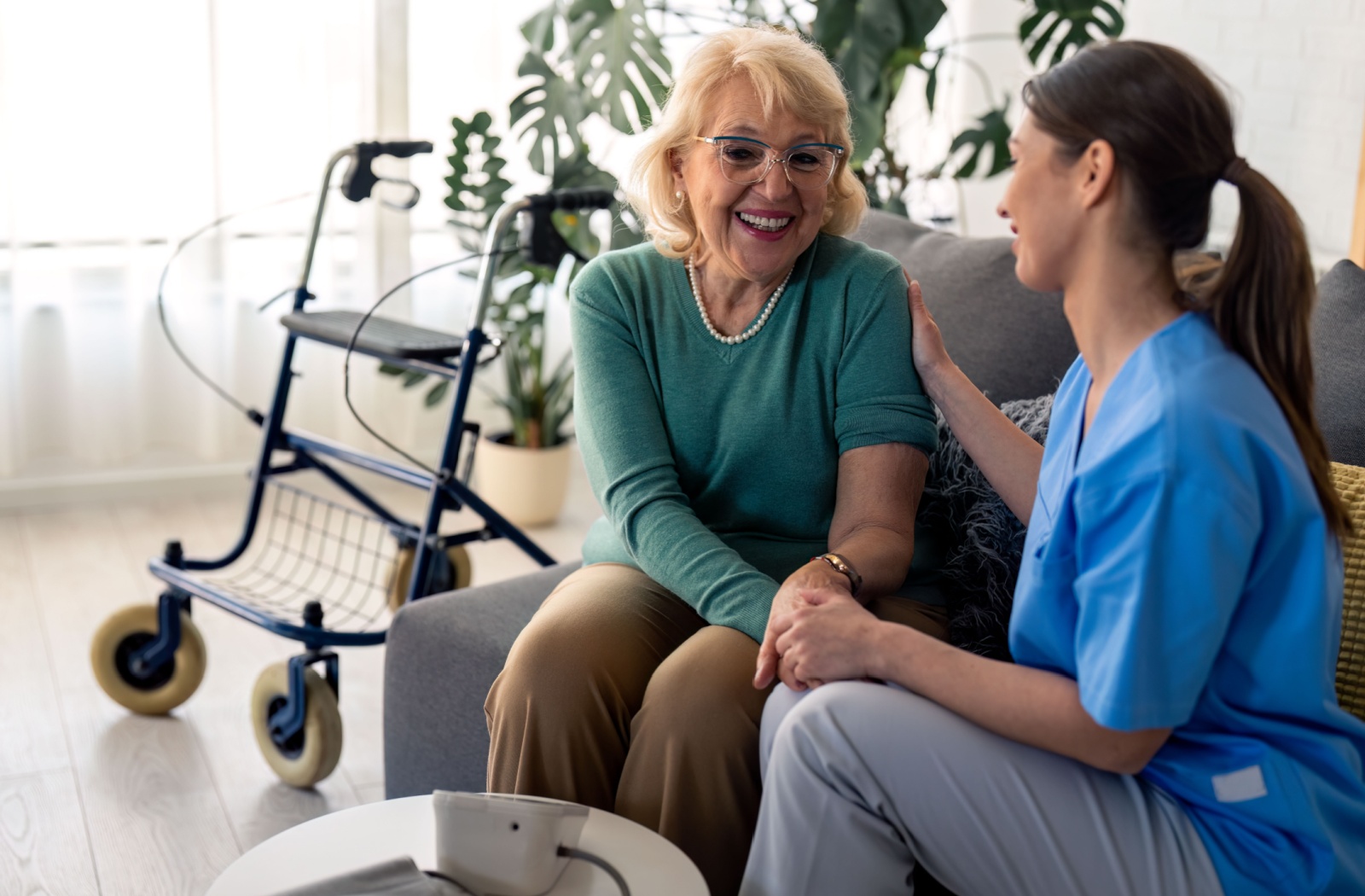 An older woman and a female nurse sitting on a couch holding hands and smiling.