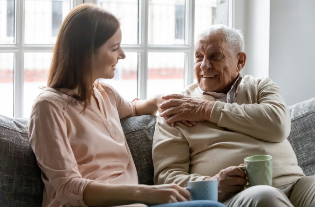 An older man and younger adult woman sitting on a couch smiling and looking at each other while holding hands.