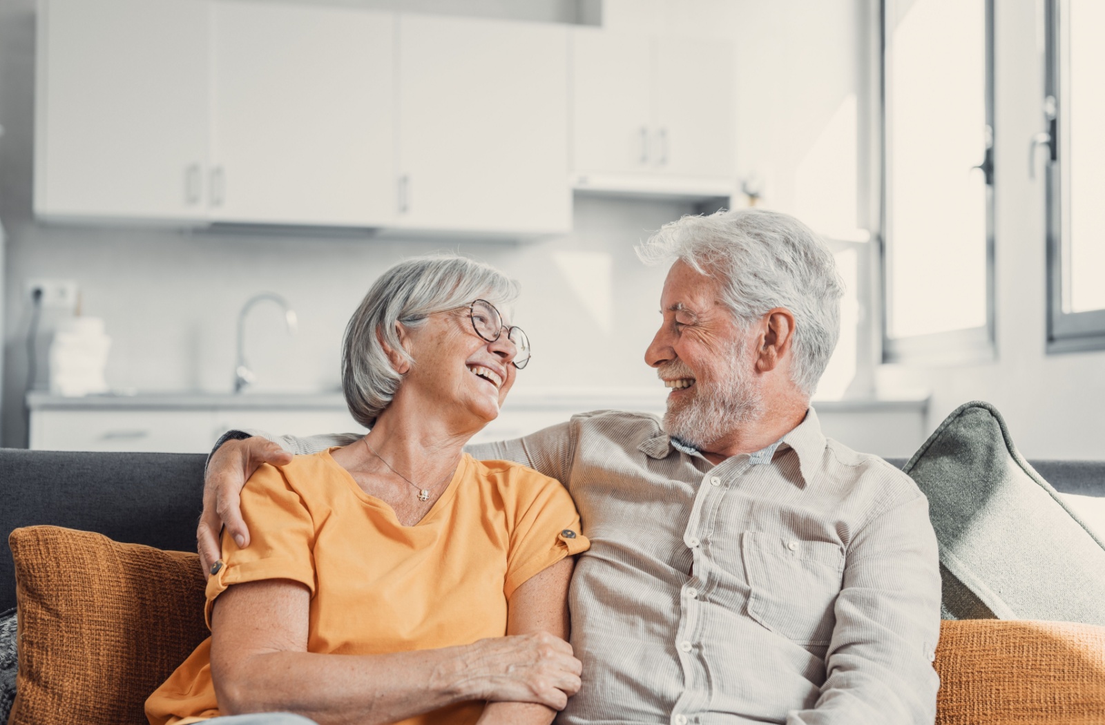 A senior couple are sitting side by side on a couch with their arms around each other - they are smiling and laughing.