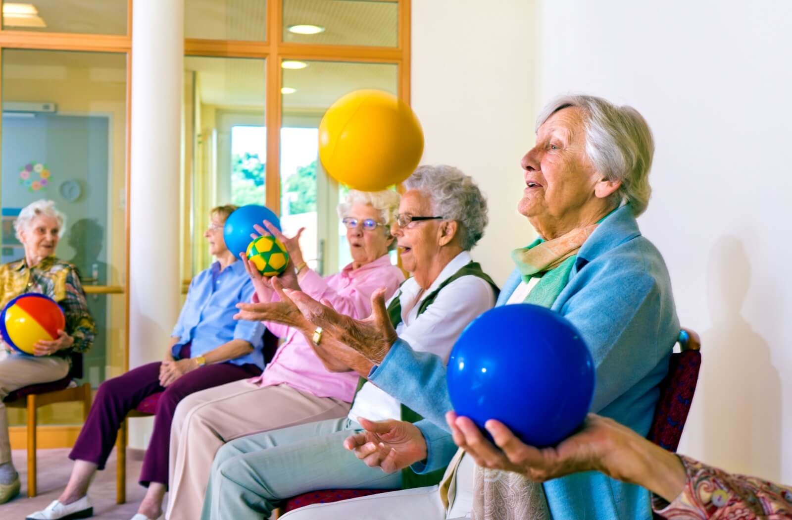 A joyful group of elderly people are sitting in chairs during an exercise class while they are tossing and catching balls.