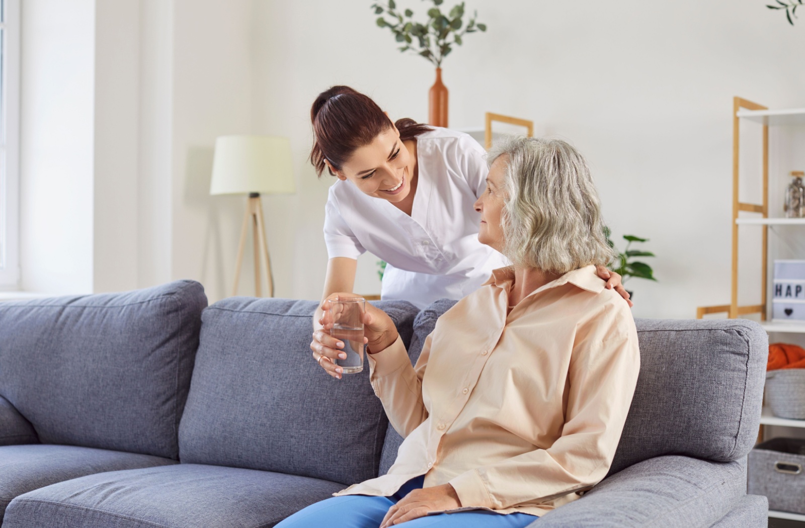 A compassionate caregiver offering water to a senior woman sitting on a couch promoting the benefits of assisted living support