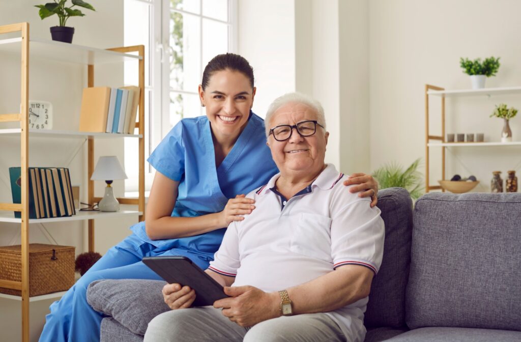 A caregiver and resident in assisted living sitting on a couch with a tablet and smiling.