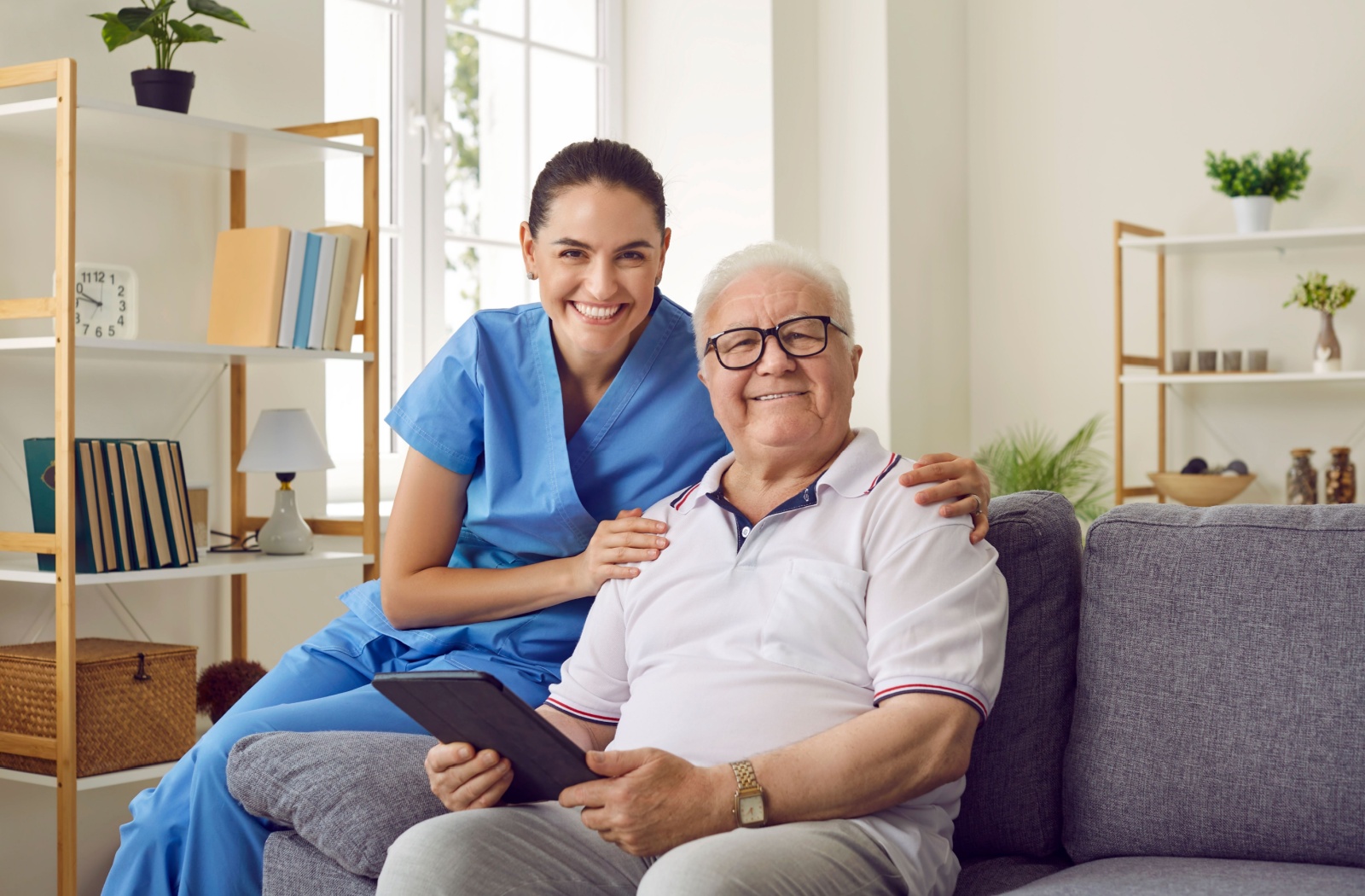 A caregiver and resident in assisted living sitting on a couch with a tablet and smiling.