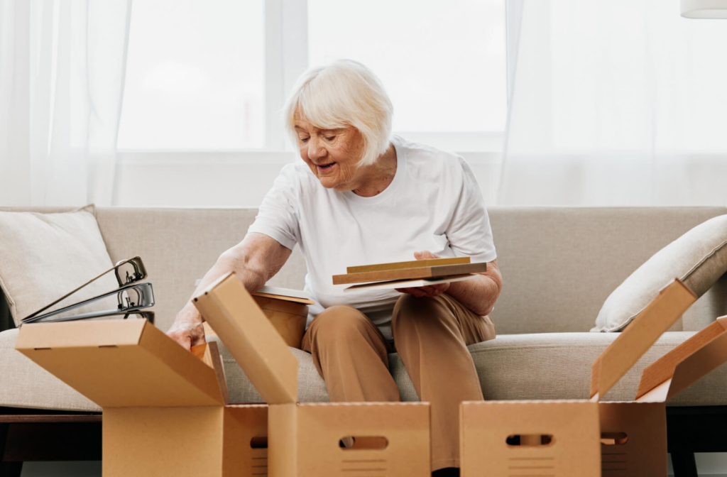 An older adult smiles while sorting between three boxes on the couch while packing their home to prepare for a move to assisted living