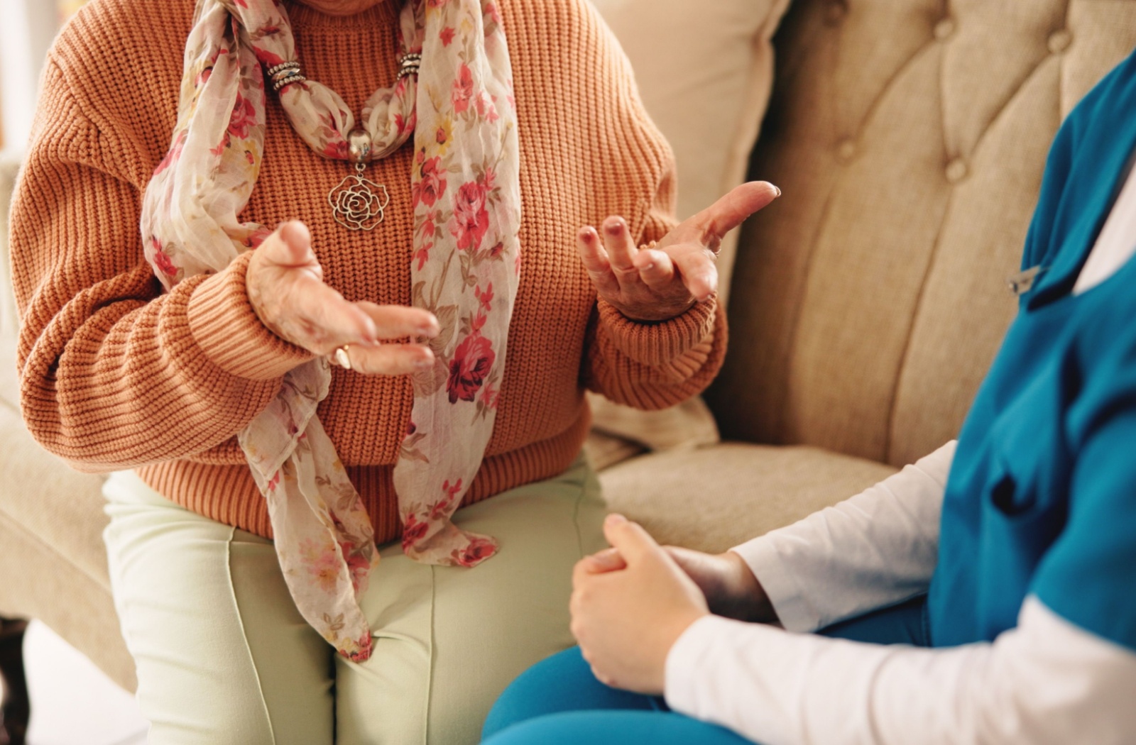A close-up image of an older adult in an orange sweater gesturing to a caregiver in assisted living while sitting on a couch